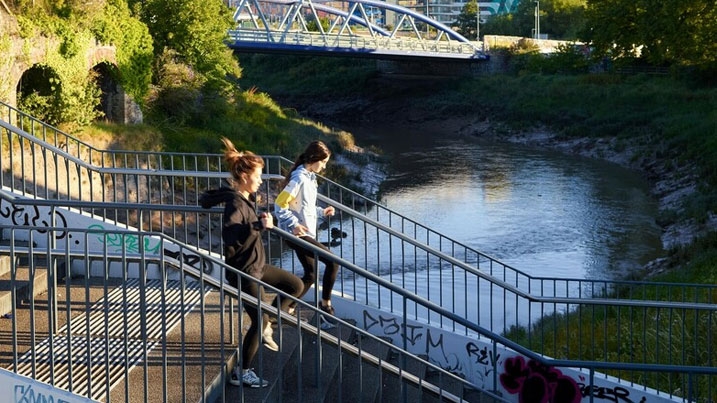 Two young women running down stairs from a bridge over an urban river