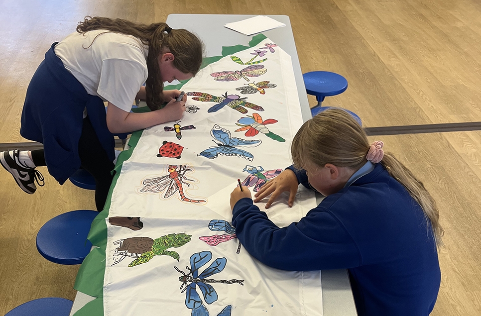 Two children concentrate while drawing and colouring dragonflies onto a white flag.
