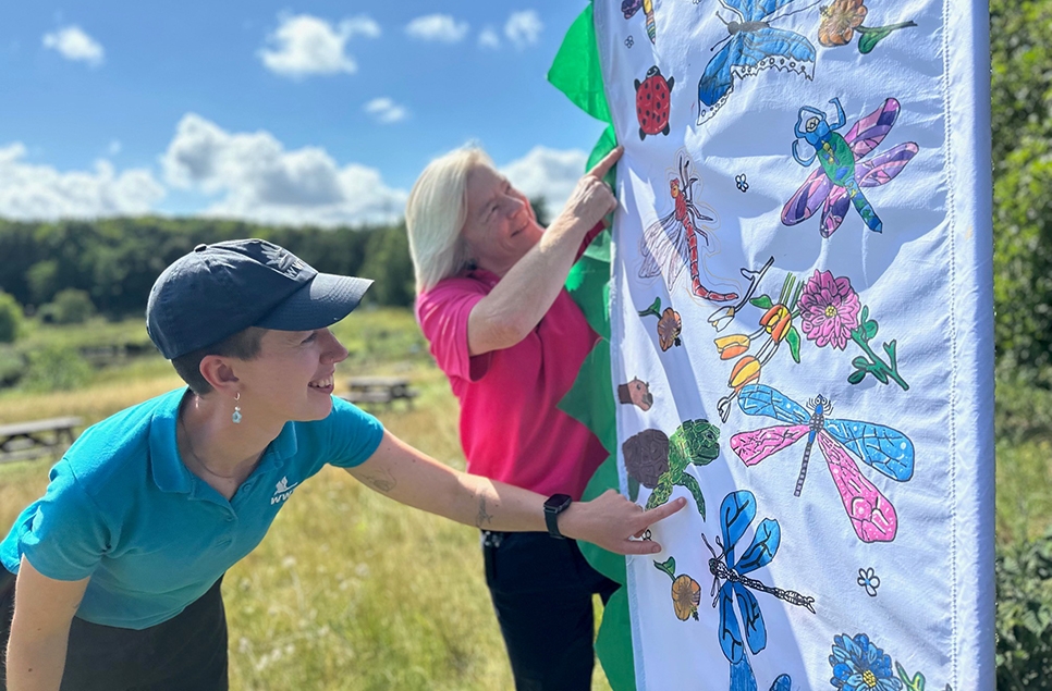 Learning Assistant Ruby Miller with Learning Manager Joanne Newbury looking at the detail of one of the flags, which has lots of blue and pink dragonfly drawings. A background of green grass and blue sky.