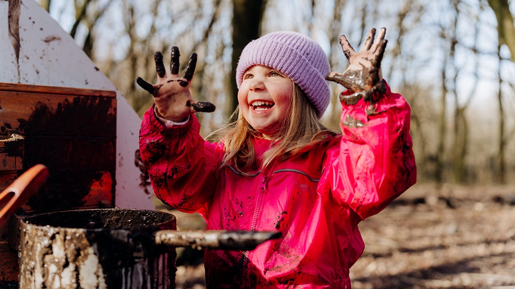 Visiting child playing in the mud kitchen, raising muddy hands up while smiling