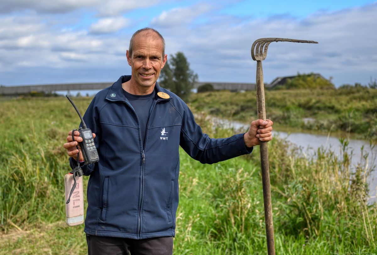 David Tough standing alongside the Hundred Foot Drain, with the WWT visitor centre roof and footbridge in the background. David is holding the rescued radio and float to the left and a long rake used in the rescue to the right. 
