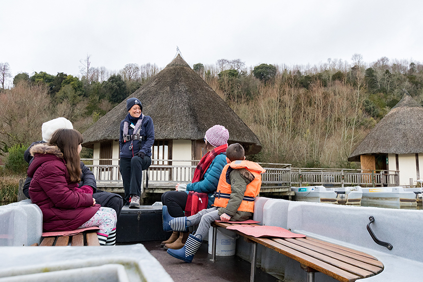 Adults and children sit in a boat talking to a member of staff of WWT with winter trees in the background