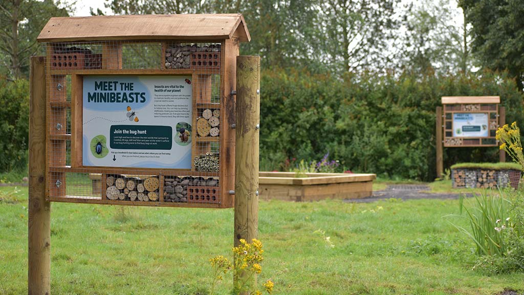 A bughouse sign at the Bug Hunting Zone at WWT Martin Mere. In the background there is a pond and a bughouse.