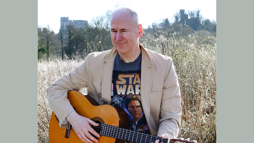 A man sitting holding a guitar with reedbeds in the background. 