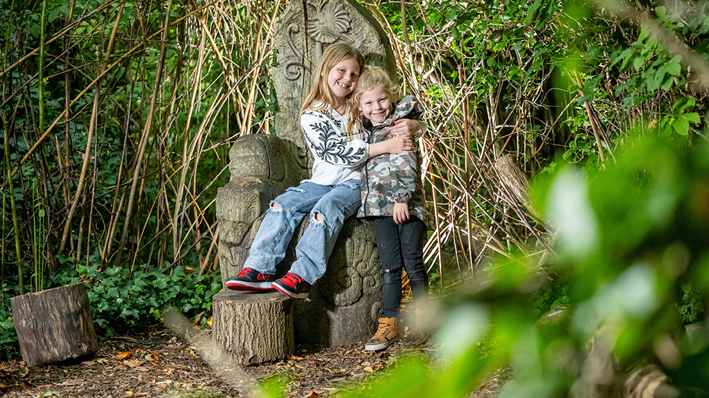 Two children sit on an upright log that has been carved into a throne.