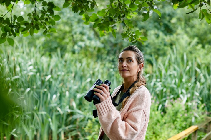 Person holding binoculars, stood outdoors and surrounded by reedbeds