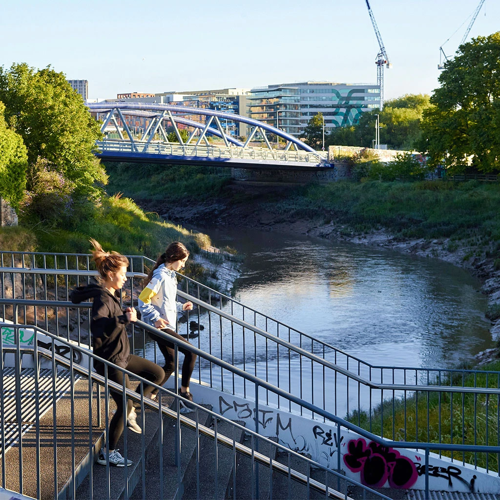 Two people running down the steps of a bridge crossing the river in Bristol. There is another bridge and buildings in the background