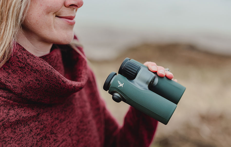 Close-up image of somebody smiling, holding a pair of dark green Swarovski binoculars