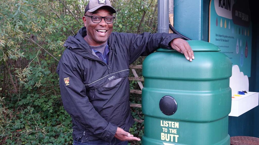 David Lindo smiles at the camera while standing next to and pointing at a water butt wihch features the words 'listen to the butt'
